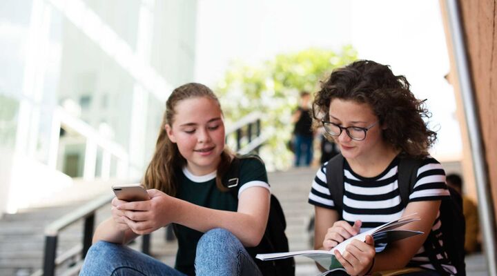 Teenage Girls Sitting On School Steps Holding Phone And Book