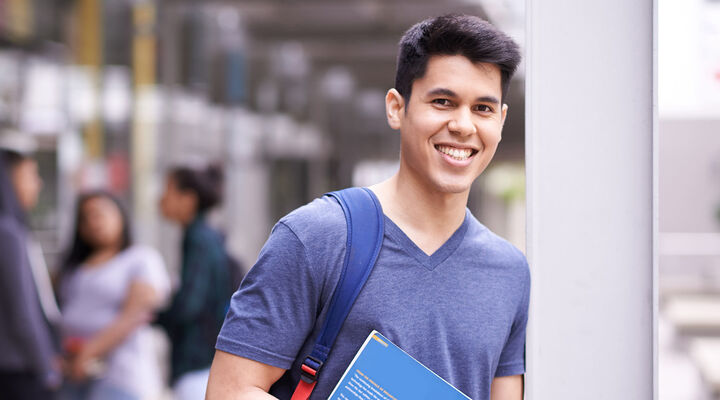 Young Man With Dark Hair In Blue Tshirt Wearing Backpack And Holding Textbook