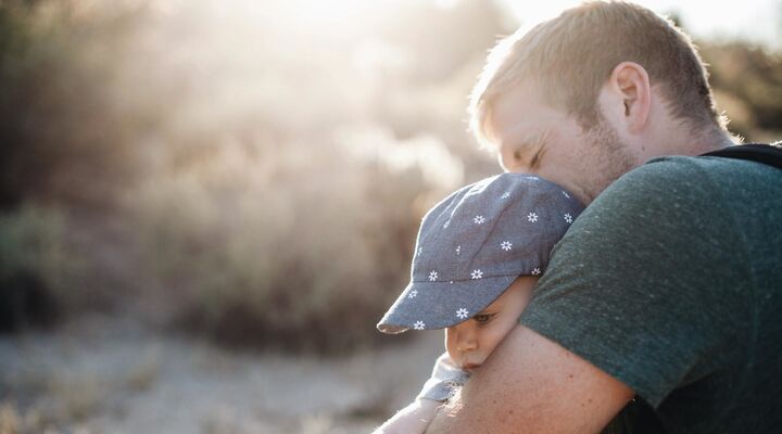 Man In Grey Tshirt Hugging Toddler In Blue Hat With White Flowers Backlit