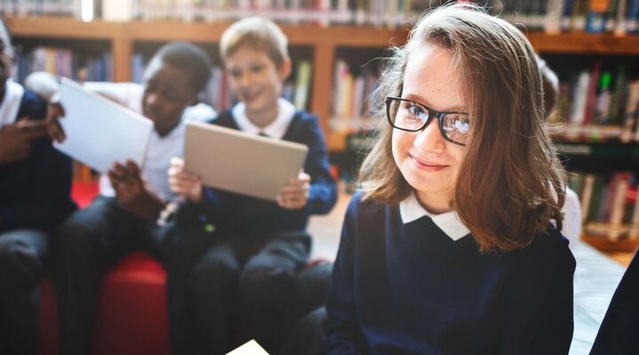 Primary School Girl In Uniform Sitting In Library Smiling At Camera With Reflection In Glasses