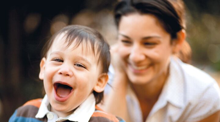 Toddler Smiling At Camera With Mother Blurred Behind Him