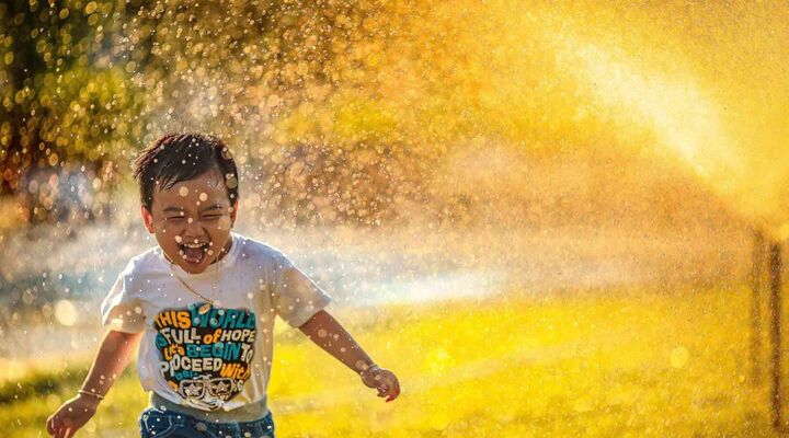 Boy Running Through Sprinklers Which Are Reflecting Bright Golden Light