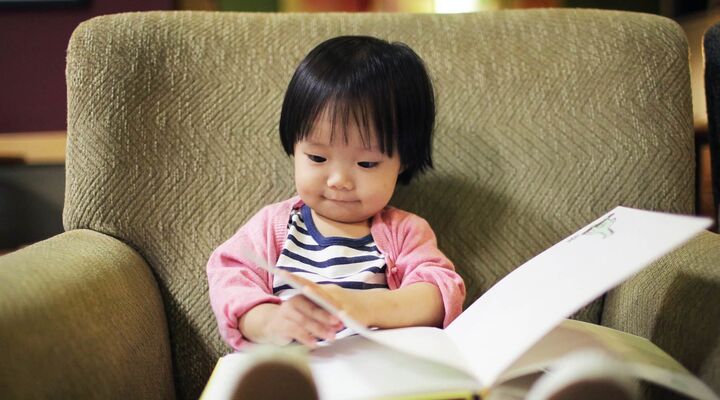 Small Girl Reading A Book In A Beige Armchair