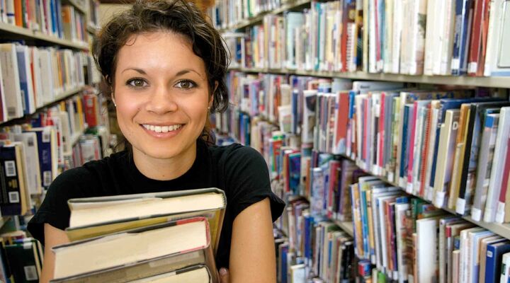 Young Woman Carrying Stack Of Books Standing In Library Rows
