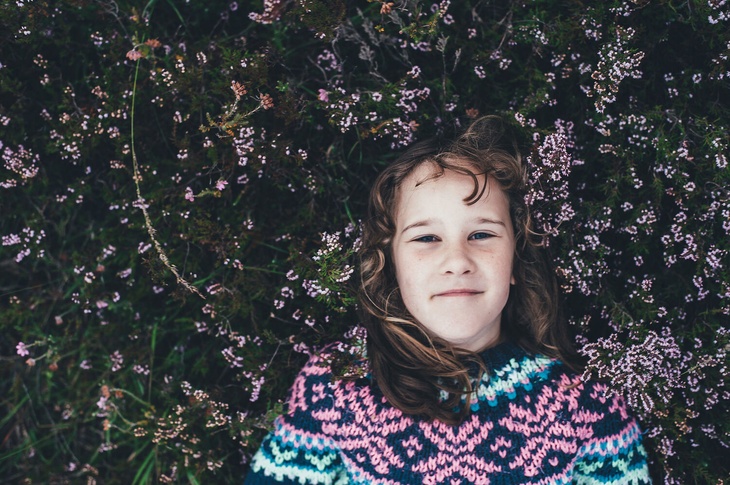 girl in colourful woollen jumper leaning against hedge with flowers