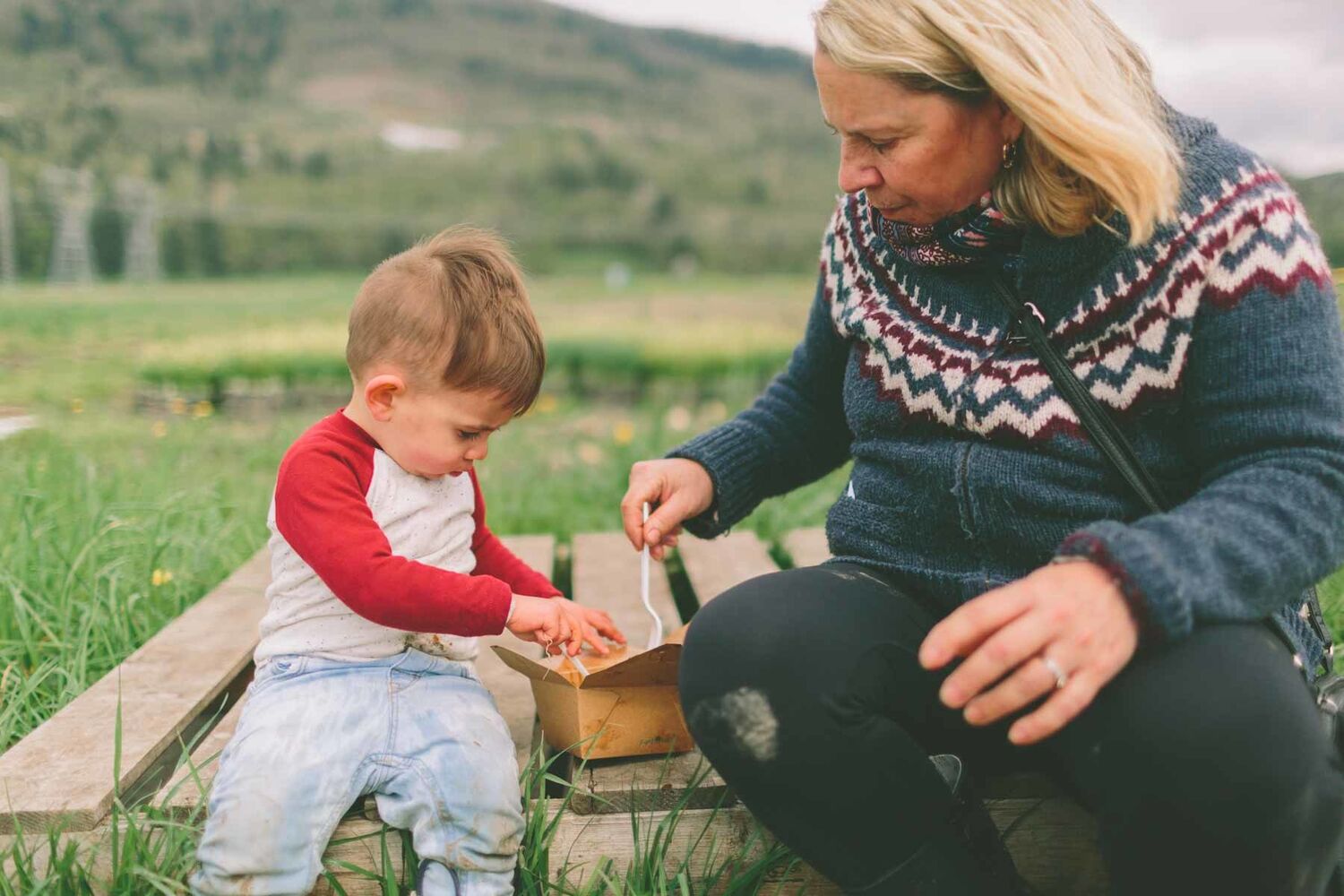 Grandmother And Toddler Sitting On Wooden Crate In Field Eating Take Away
