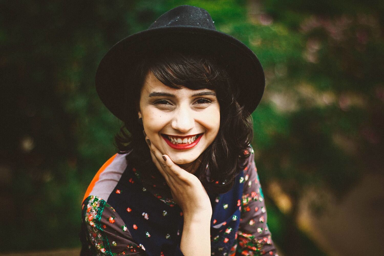 Young Woman With Red Lipstick Wearing Black Felt Hat Smiling At Camera With Greenery Behind Her