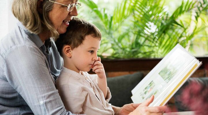Boy sitting on grandmothers lap whilst reading book on couch with ferns in background