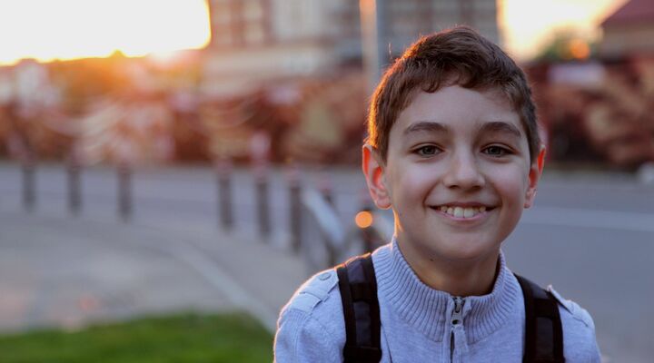 Boy Wearing Backpack Smiling With Sunset And Buildings Behind Him