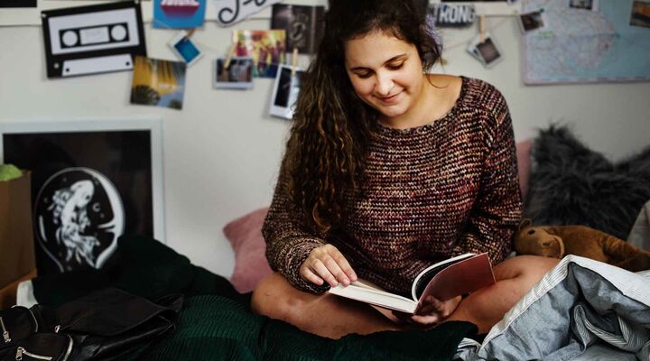 Teenage Girl In Knitted Jumper Reading Book On Her Bed With Pictures On Wall