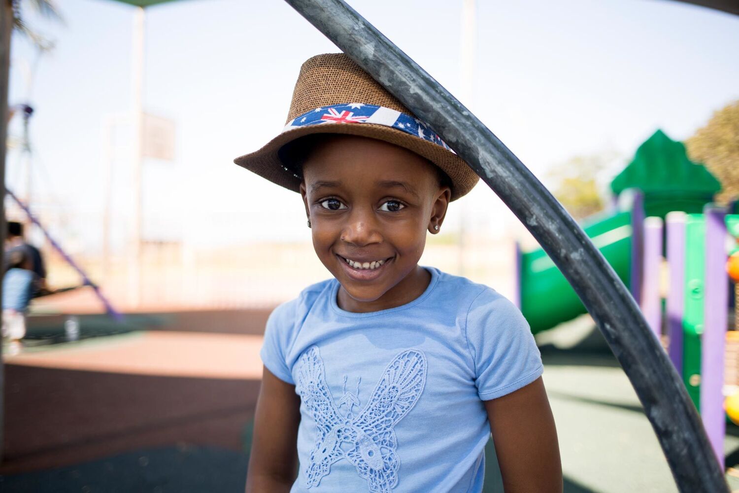 Young Aboriginal Girl In Blue Shirt And Hat Standing In A Park Smiling