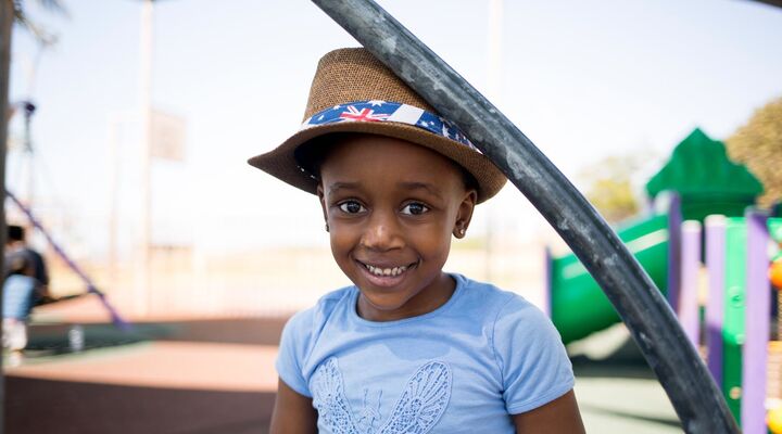Young Aboriginal Girl In Blue Shirt And Hat Standing In A Park Smiling