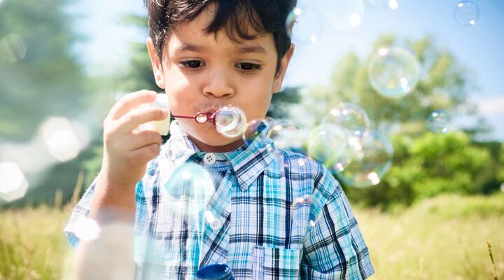Aboriginal Boy In Blue Checked Shirt Blowing Bubbles In Field