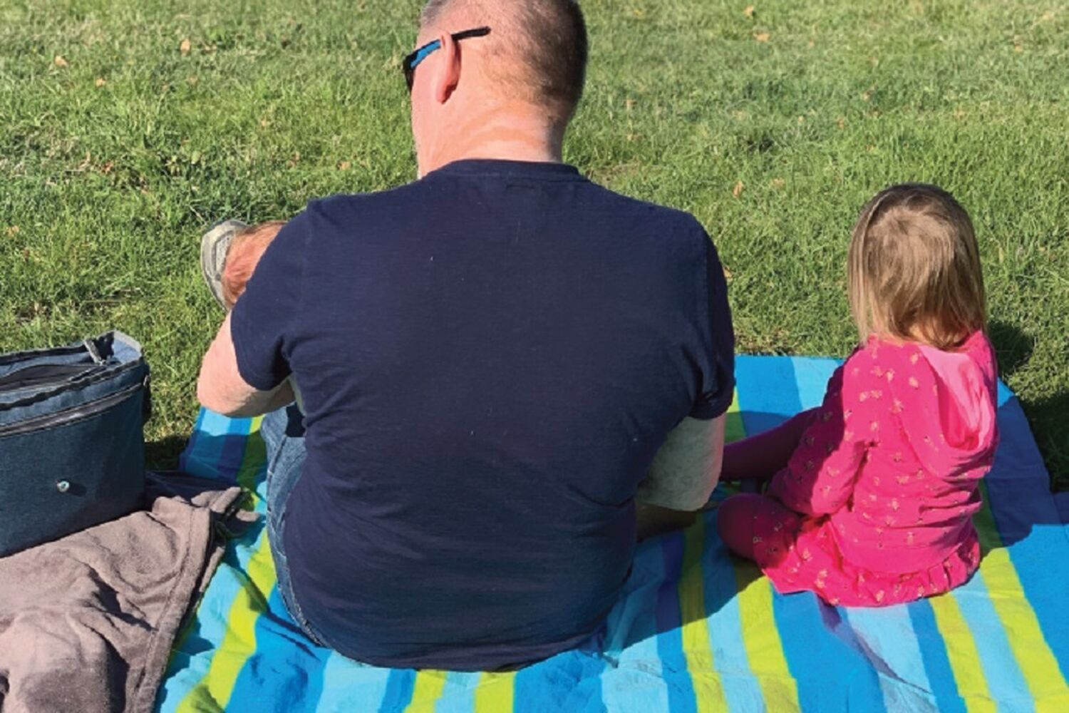 Dad Sitting With Baby In Arms And Child On A Picnic Rug