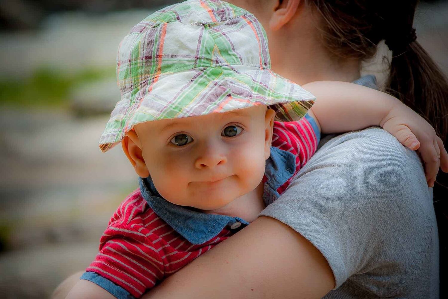 Baby In Red Polo Top And Tartan Hat Looking Over Womans Shoulder