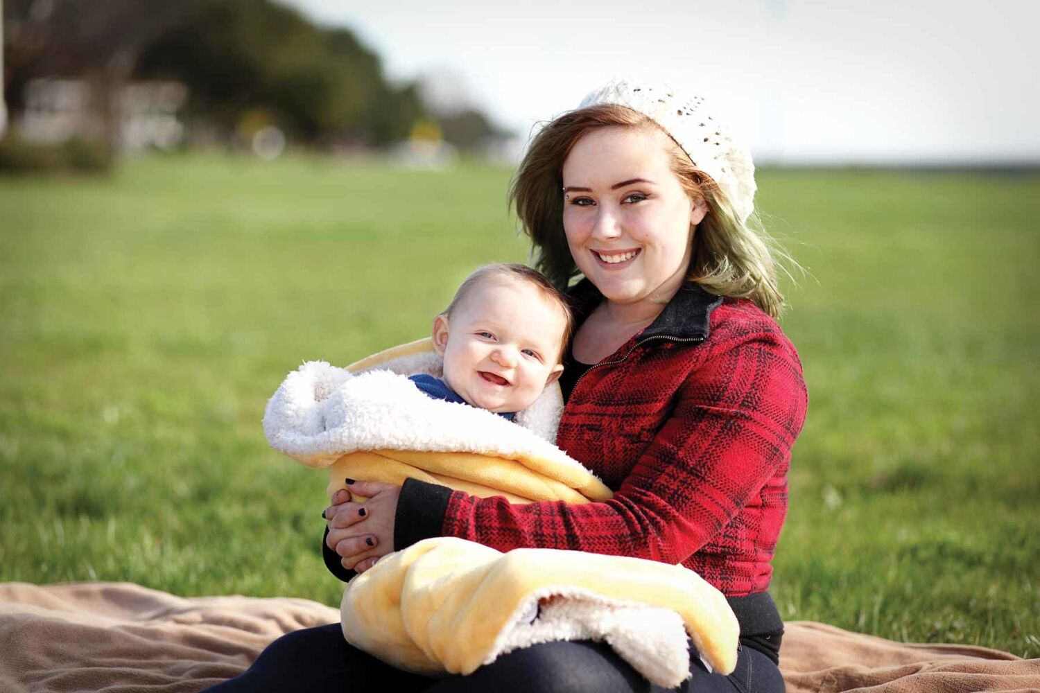 Young Mother In Tartan Jacket Holding Baby In Fleece Run Sitting On Blanket In Grass