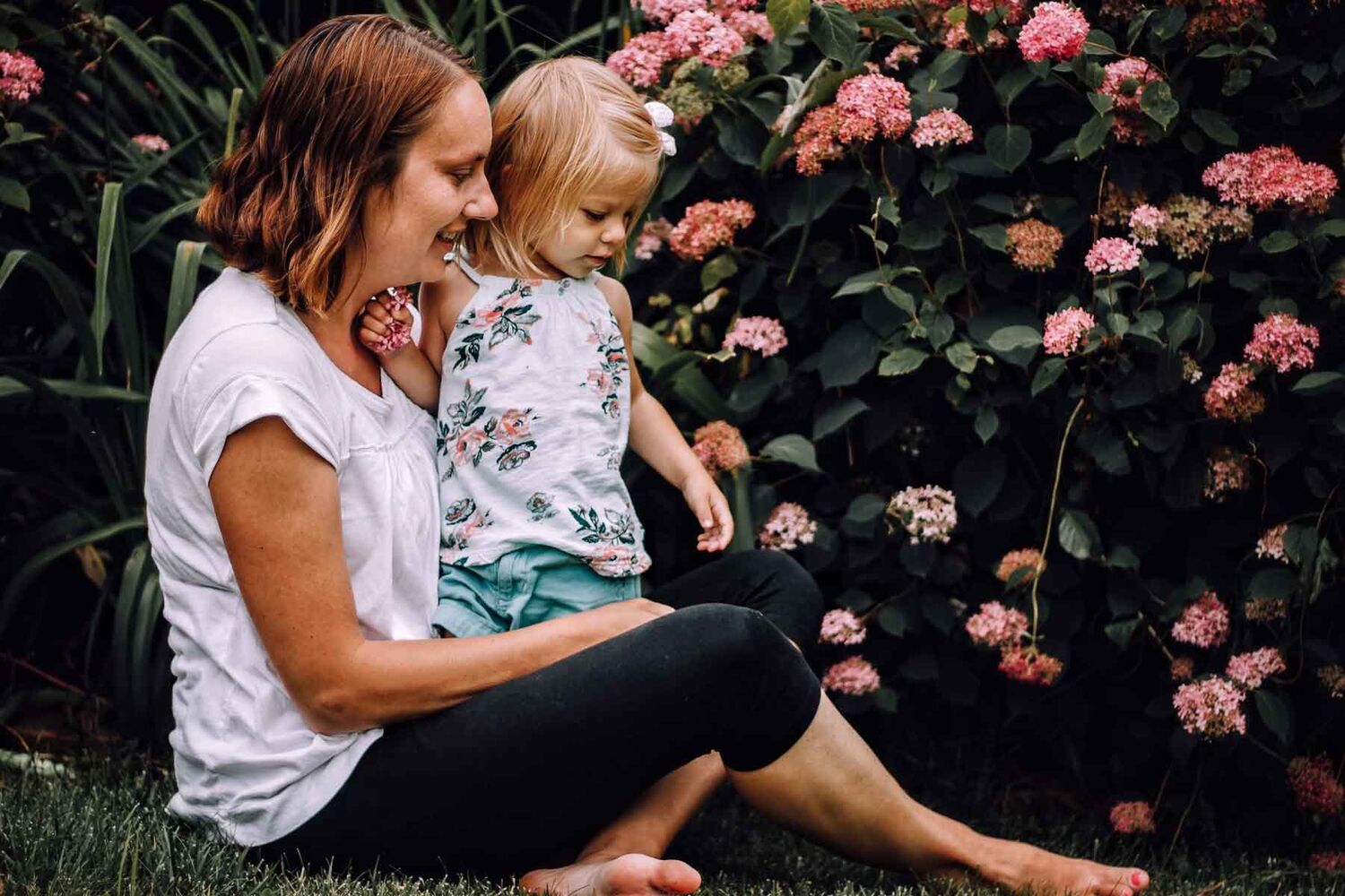 Mother Sitting On Grass With Daughter Standing In Lap In Garden