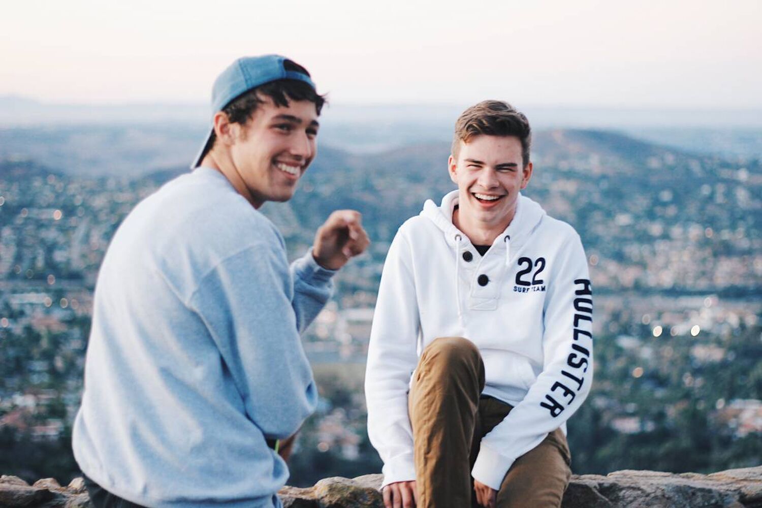 Teenage Boys In Sweatshirts Laughing Sitting In Ledge Above Town In Background