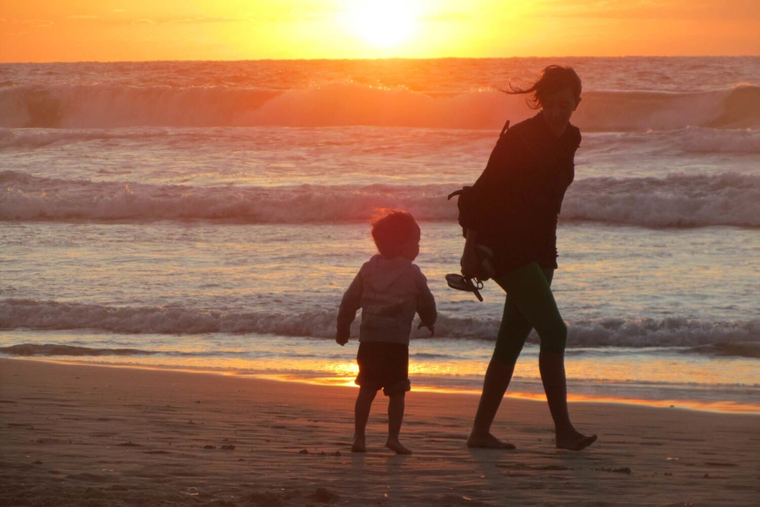 Silhouette Of Mother And Young Boy Walking On The Beach At Sunset