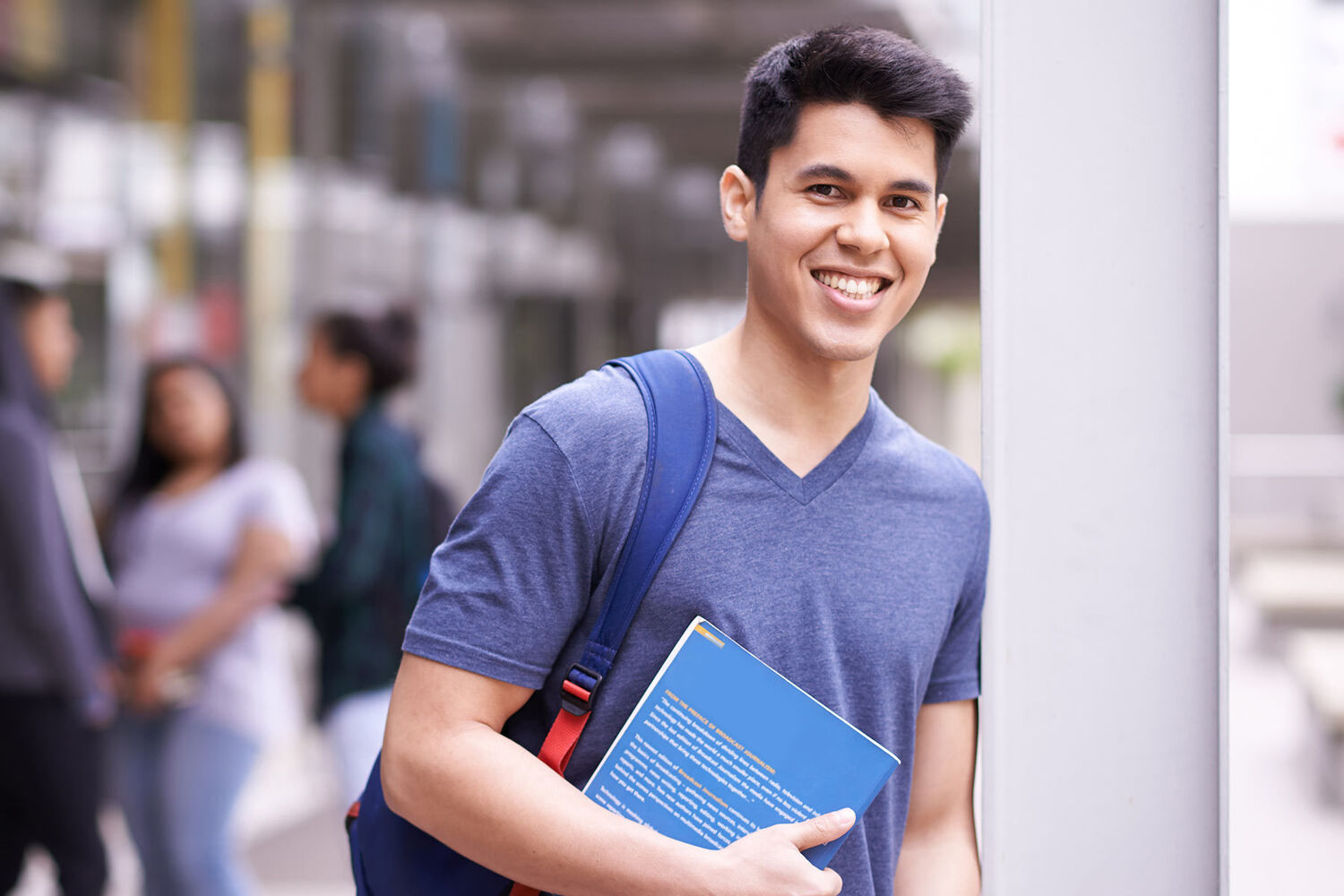 Young Man With Dark Hair In Blue Tshirt Wearing Backpack And Holding Textbook